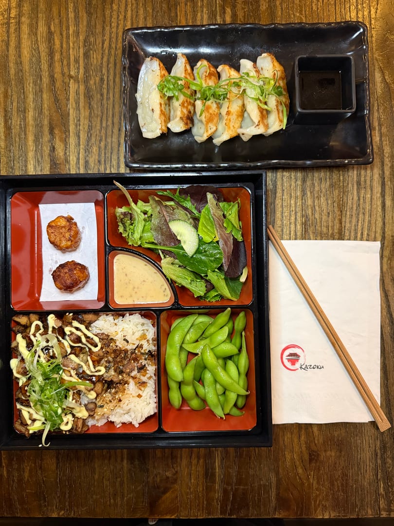 A Kazoku bento box with chicken, rice, edamame, salad, fried scallops, and gyoza.