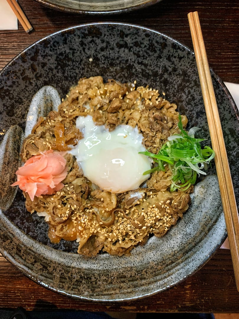 A close-up of a Gyudon bowl with beef, a poached egg, pickled ginger, and green onions.