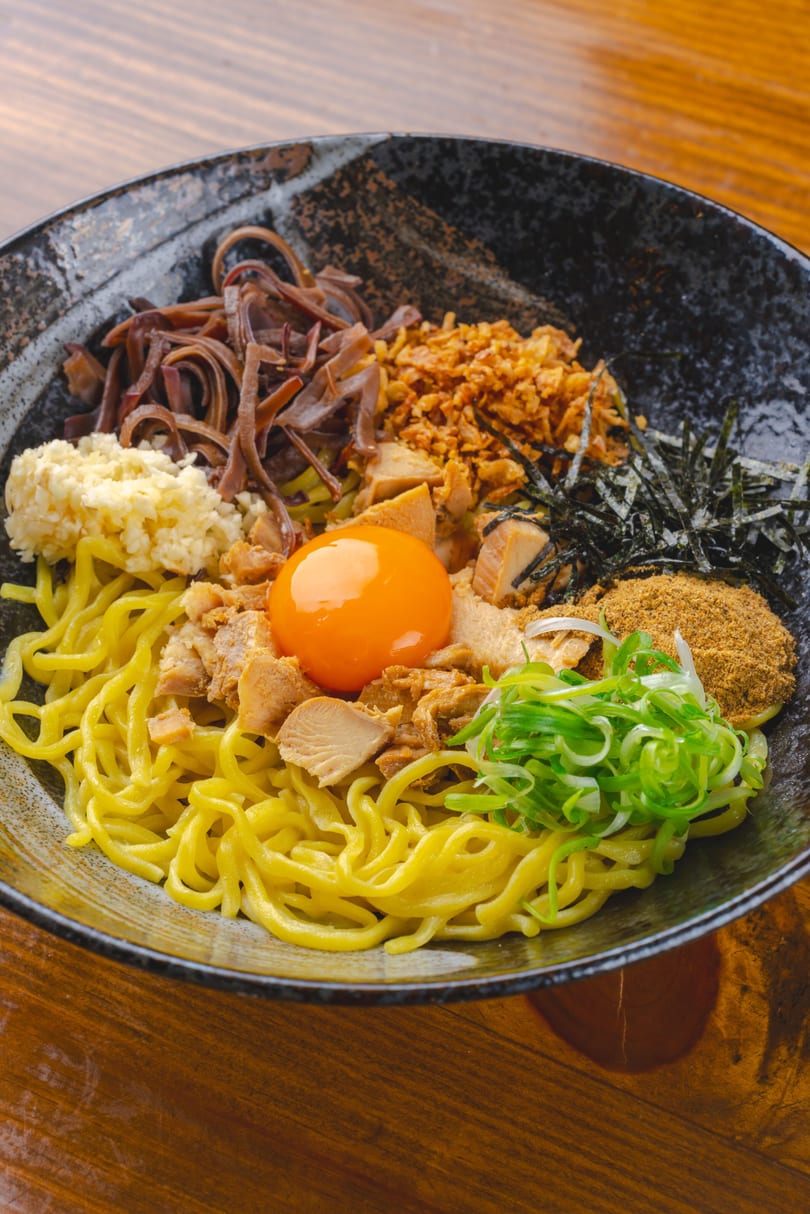 A bowl of mazesoba with noodles, pork, egg yolk, green onions, and other toppings.