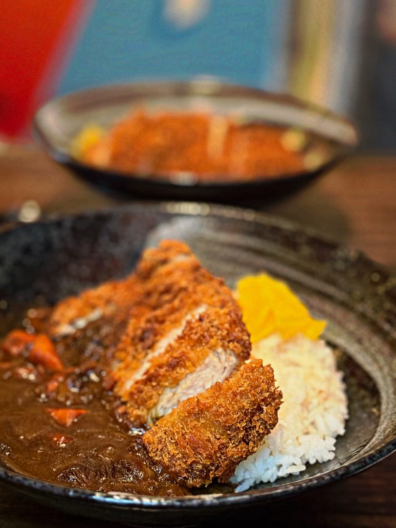 Crispy tonkatsu cutlet with rich curry sauce, white rice, and yellow pickled daikon in a black bowl.