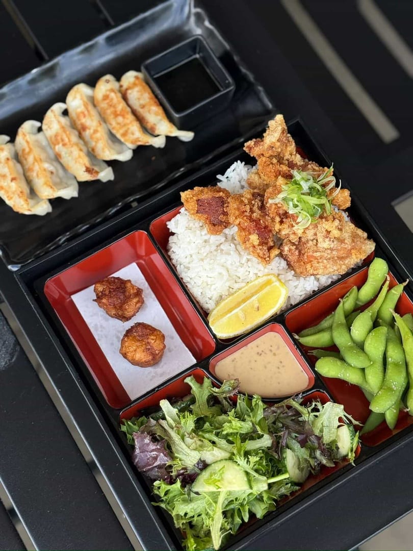 A bento box with fried chicken, rice, lemon, edamame, salad, dipping sauce, and gyoza.